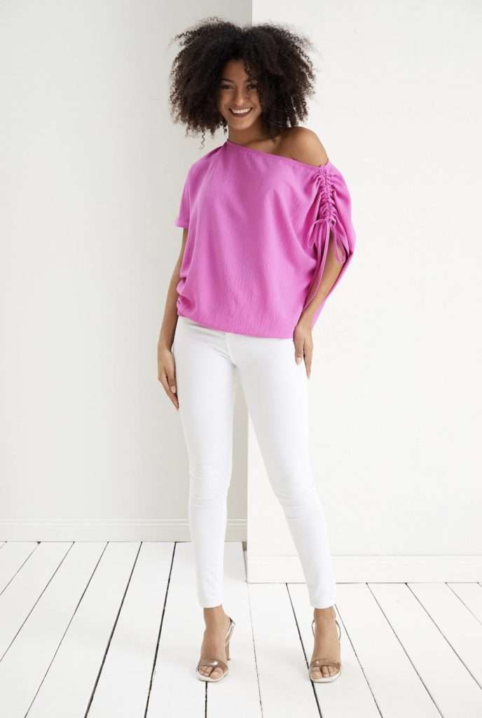 Oversized off the shoulder top shown in pink and worn with white jeans. Adjustable shoulder details